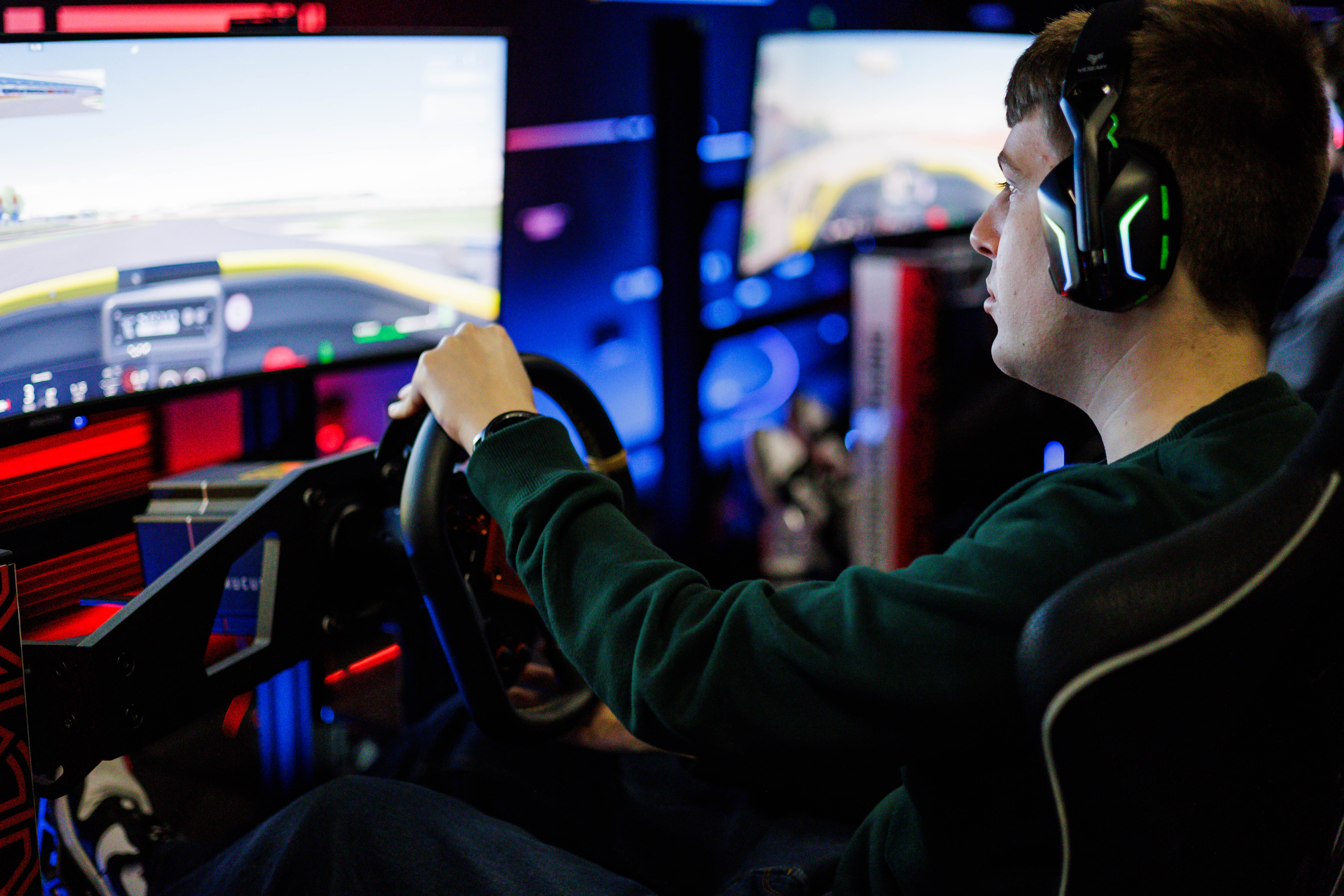 A man sat with headphones on in a professional simulator rig, looking at the wide-screen with his hands on the steering wheel and feet on the pedals.